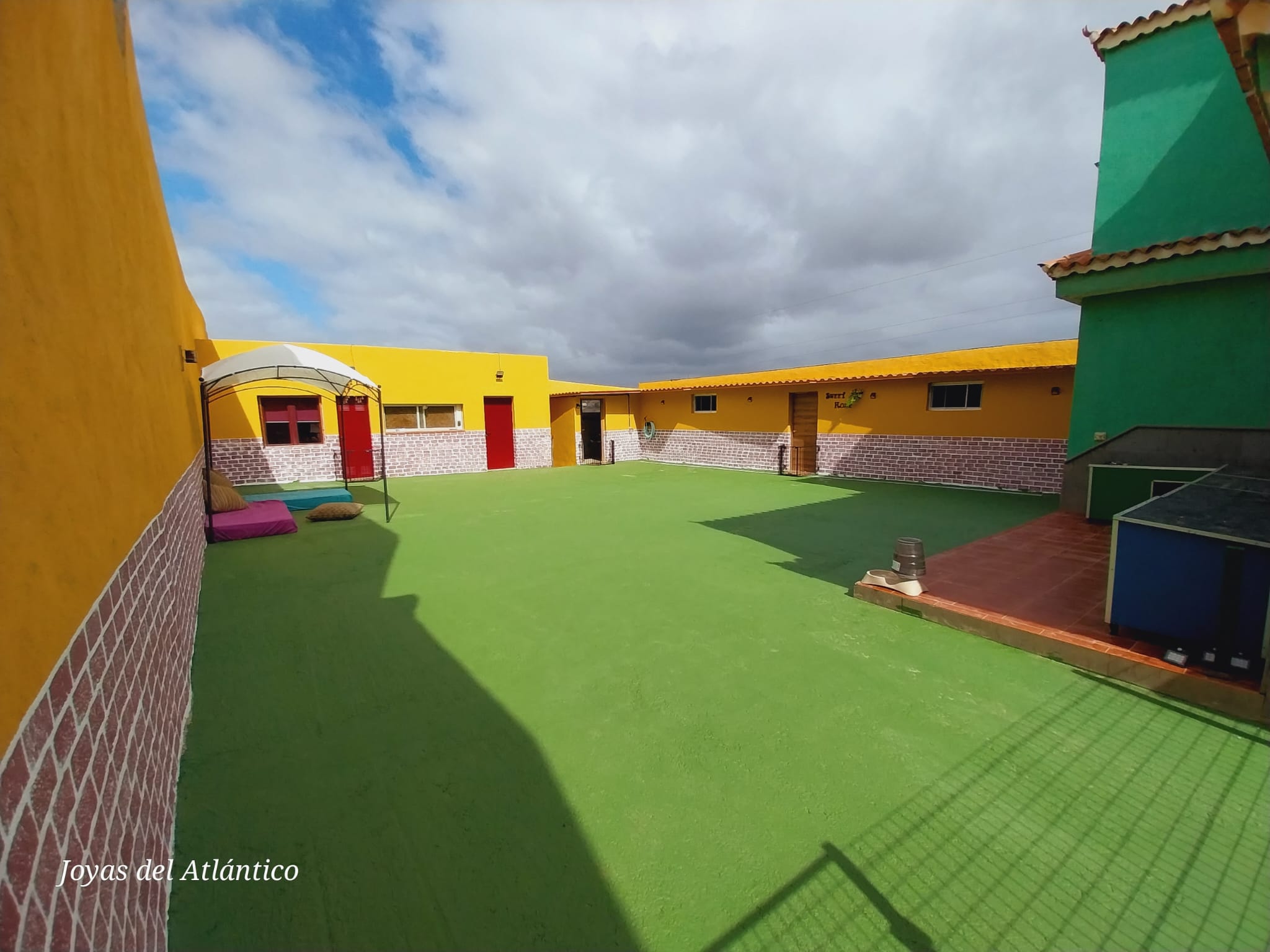 Espacio exterior de Joyas del Atlántico, con césped verde y paredes amarillas y verdes. Se observa un área de descanso con cojines y sombrilla, ideal para actividades al aire libre bajo un cielo parcialmente nublado.