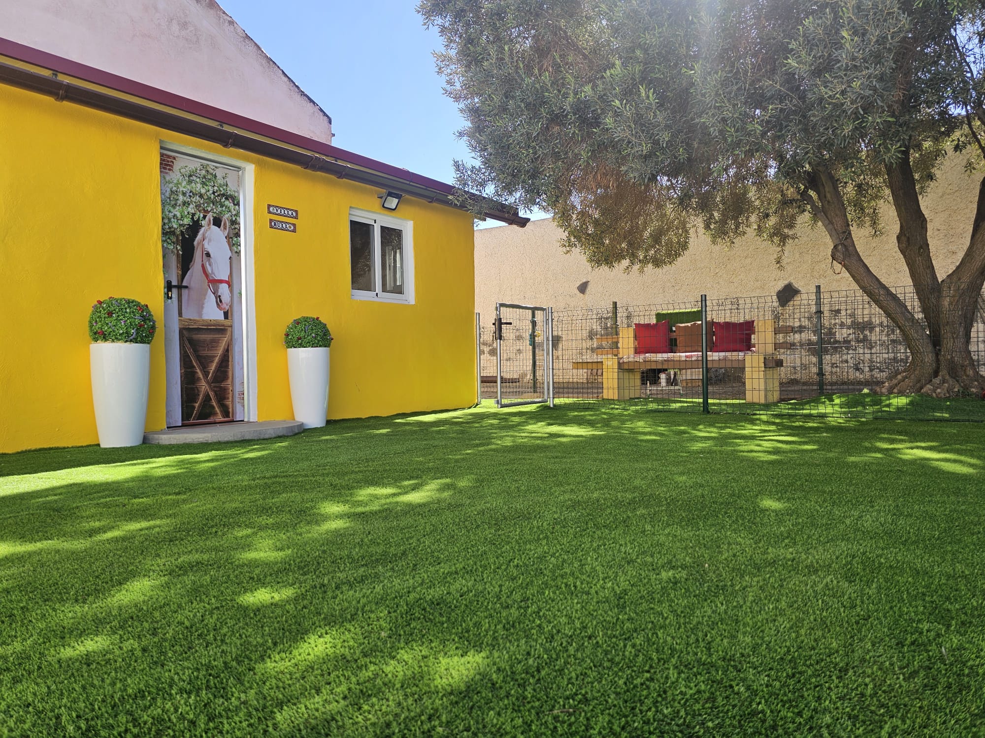 Casa amarilla con puerta decorada, rodeada de césped artificial verde, macetas blancas y un área de descanso con cojines rojos, todo en un ambiente soleado.