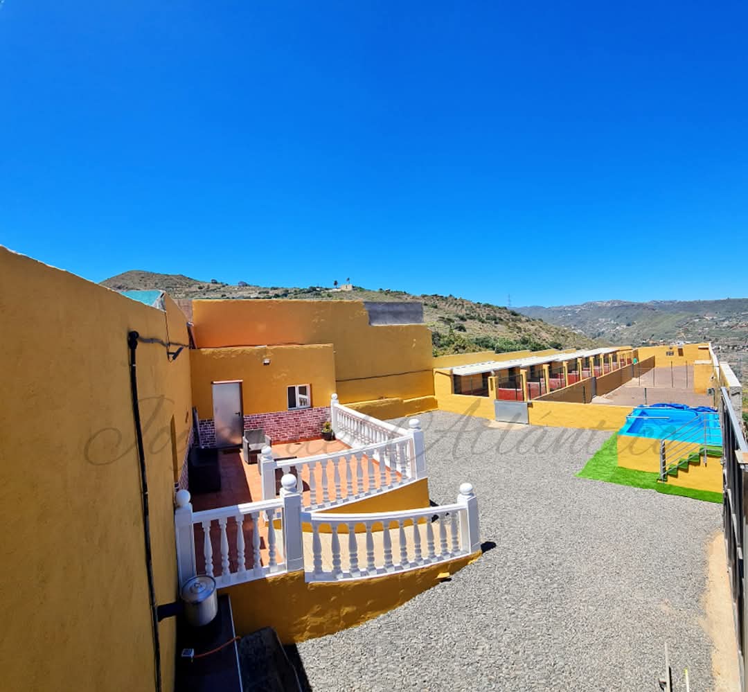 Vista panorámica de un complejo residencial con paredes amarillas, patio con barandales blancos, y área de piscina. Se aprecian montañas al fondo bajo un cielo despejado. Ideal para vacaciones y escapadas.